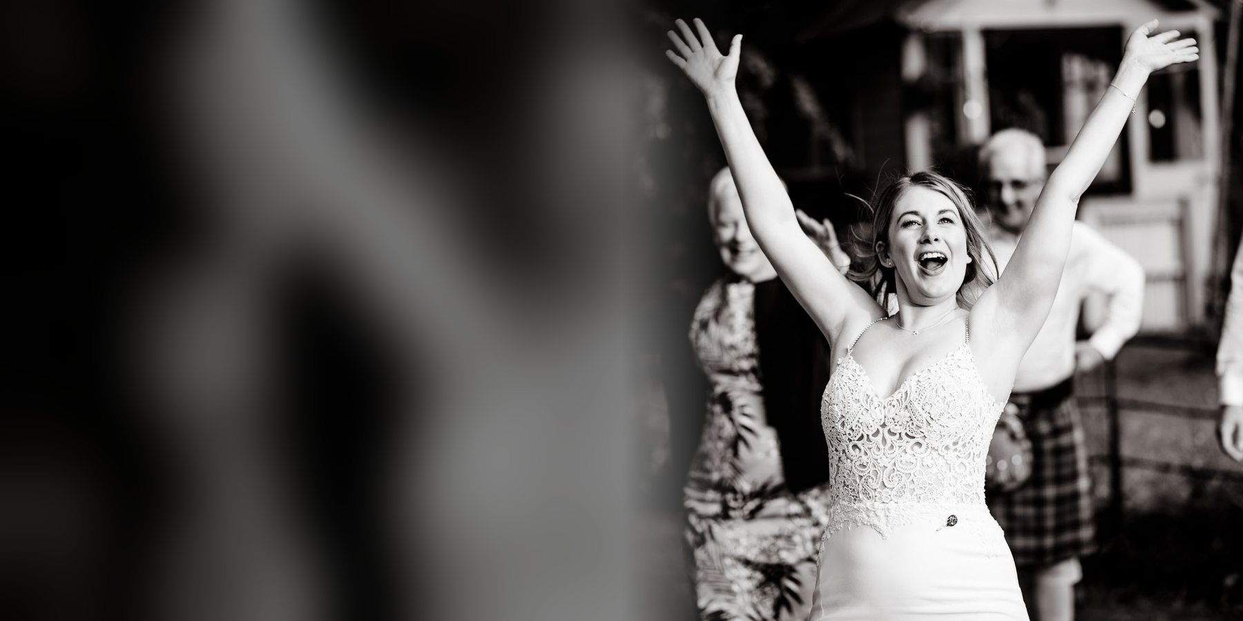 A bride throwing both arms in the air mid-celebration, guests and a kilted guest behind her